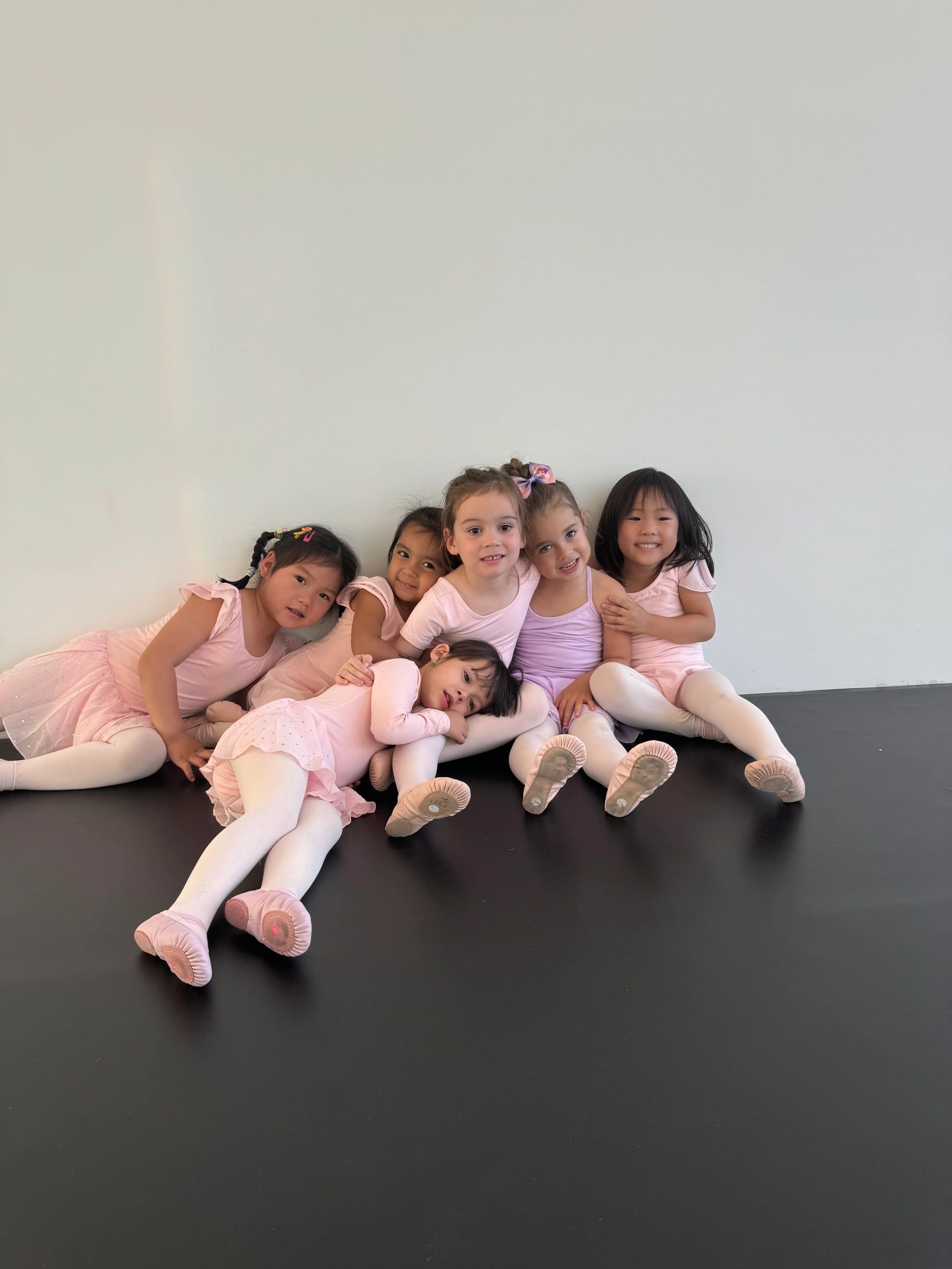 Six young dancers in pink ballet costumes resting against the studio wall.
