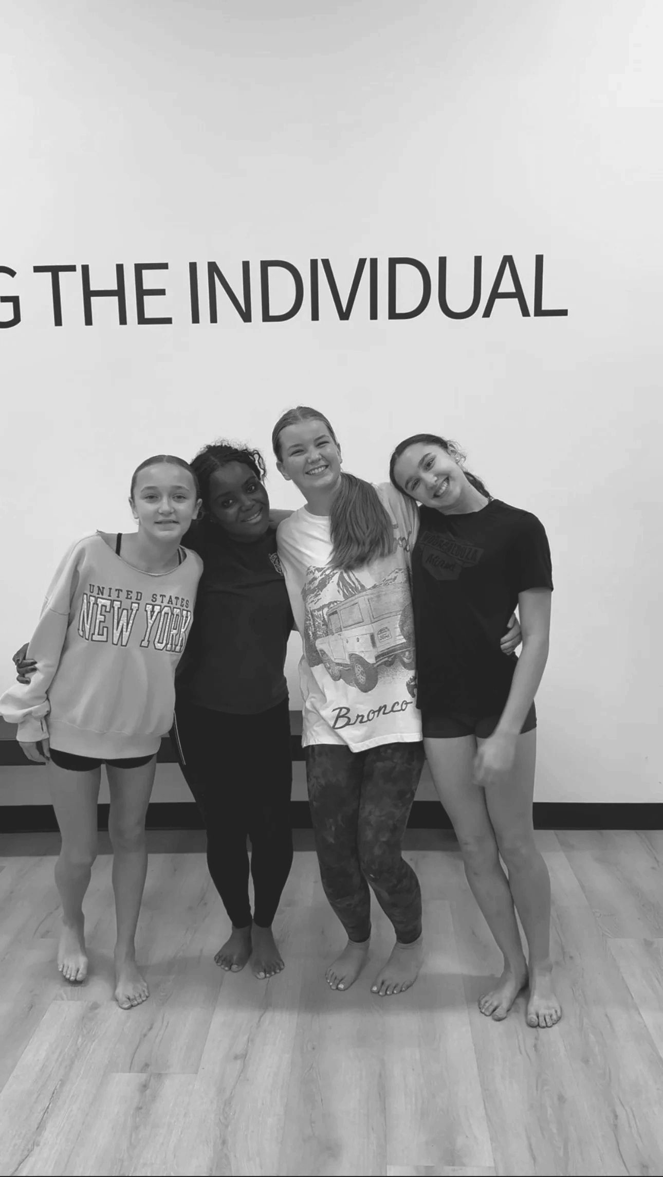 Four teen dancers smiling together in front of the &lsquo;Growing the Individual&rsquo; wall at Dance Athletics Studio.
